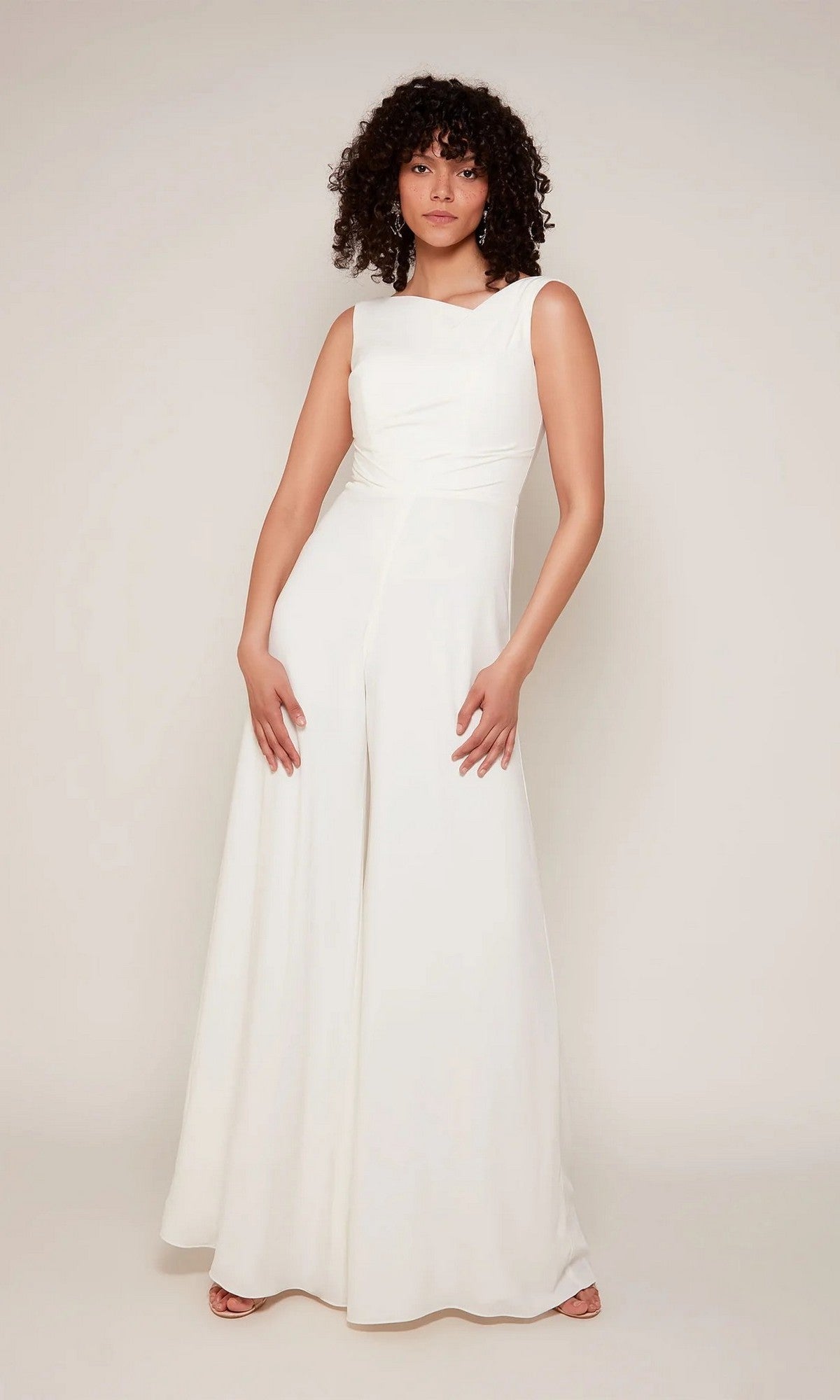 A person stands against a plain background wearing the Alyce 70052 Long White Jumpsuit in Diamond White—sleeveless and wide-leg—paired with open-toed shoes.